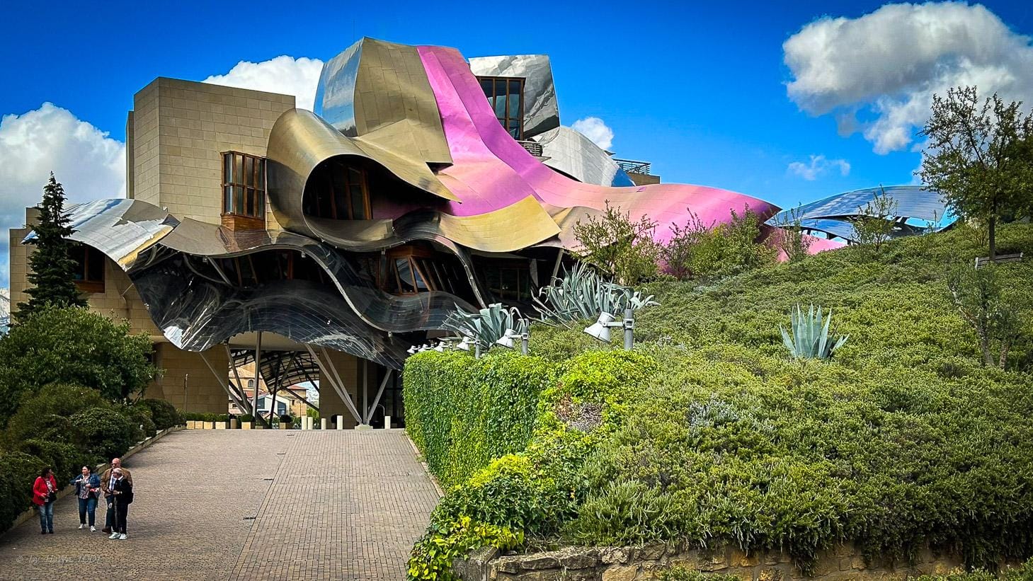 Buntes, modernes Gebäude mit wellenförmigen Formen in grüner Umgebung, sonniger Himmel.