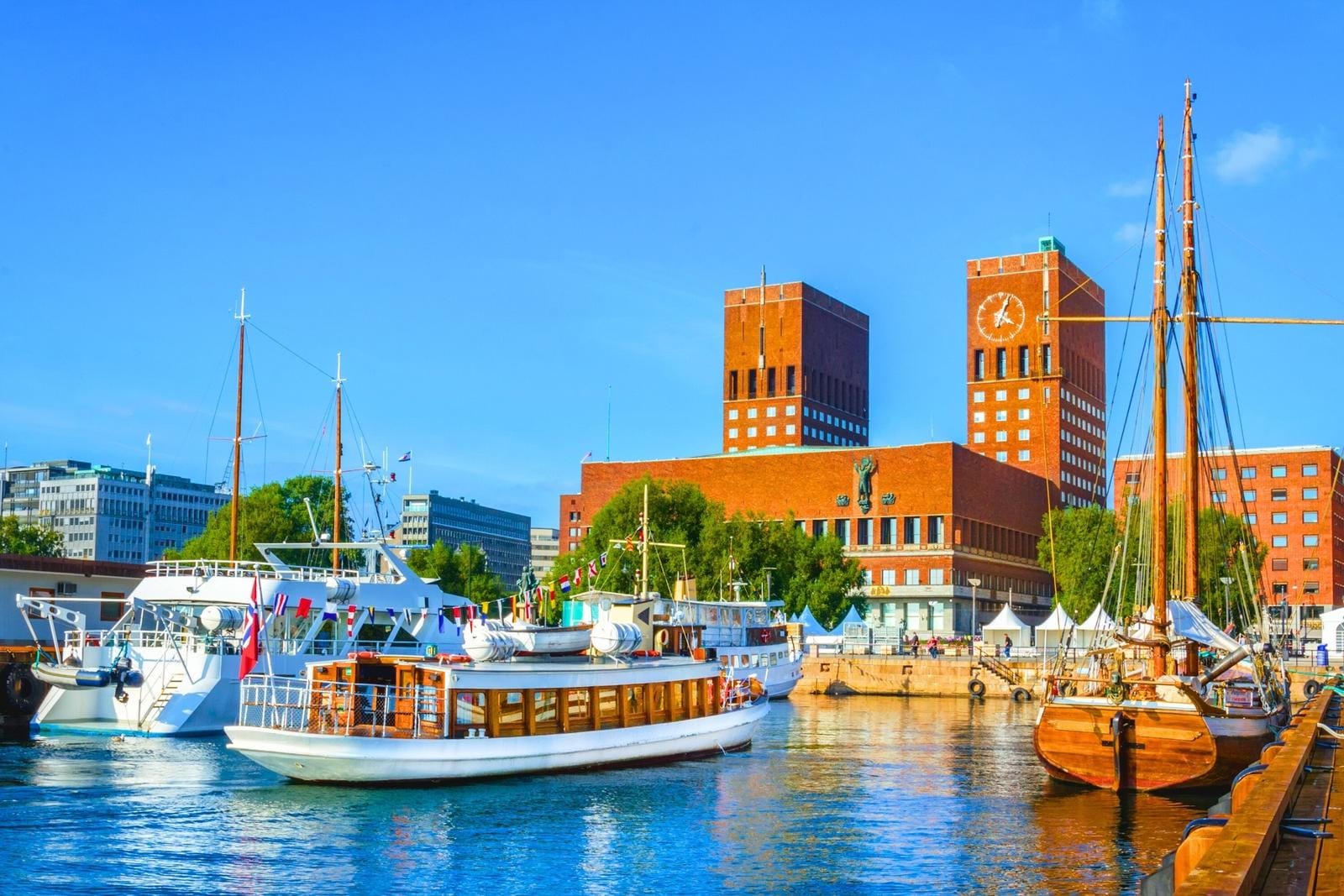 Bunte Boote im Hafen von Oslo vor dem markanten Rathaus bei strahlend blauem Himmel.