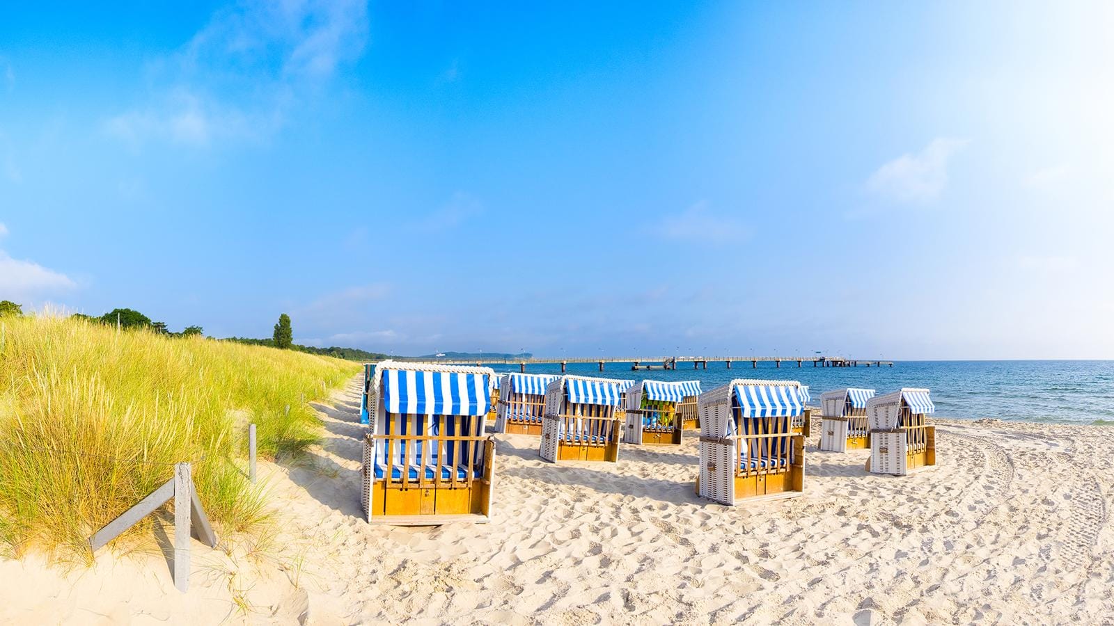 Küstenbild von Rügen mit Strandkörben, Sanddünen und einem langen Steg am Meer.