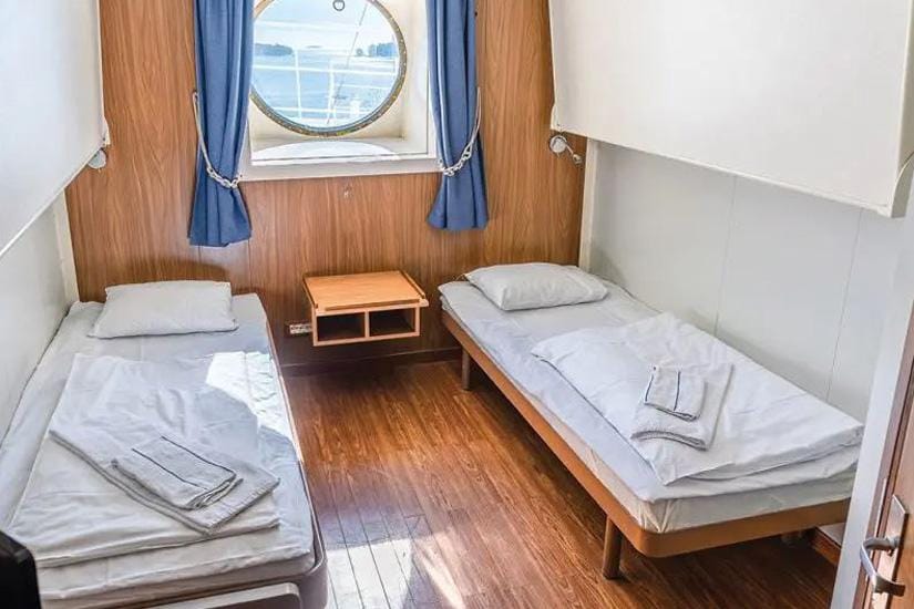 Kabine auf einem Schiff mit Bullauge und zwei Betten, vielleicht auf einer Nordseekreuzfahrt.