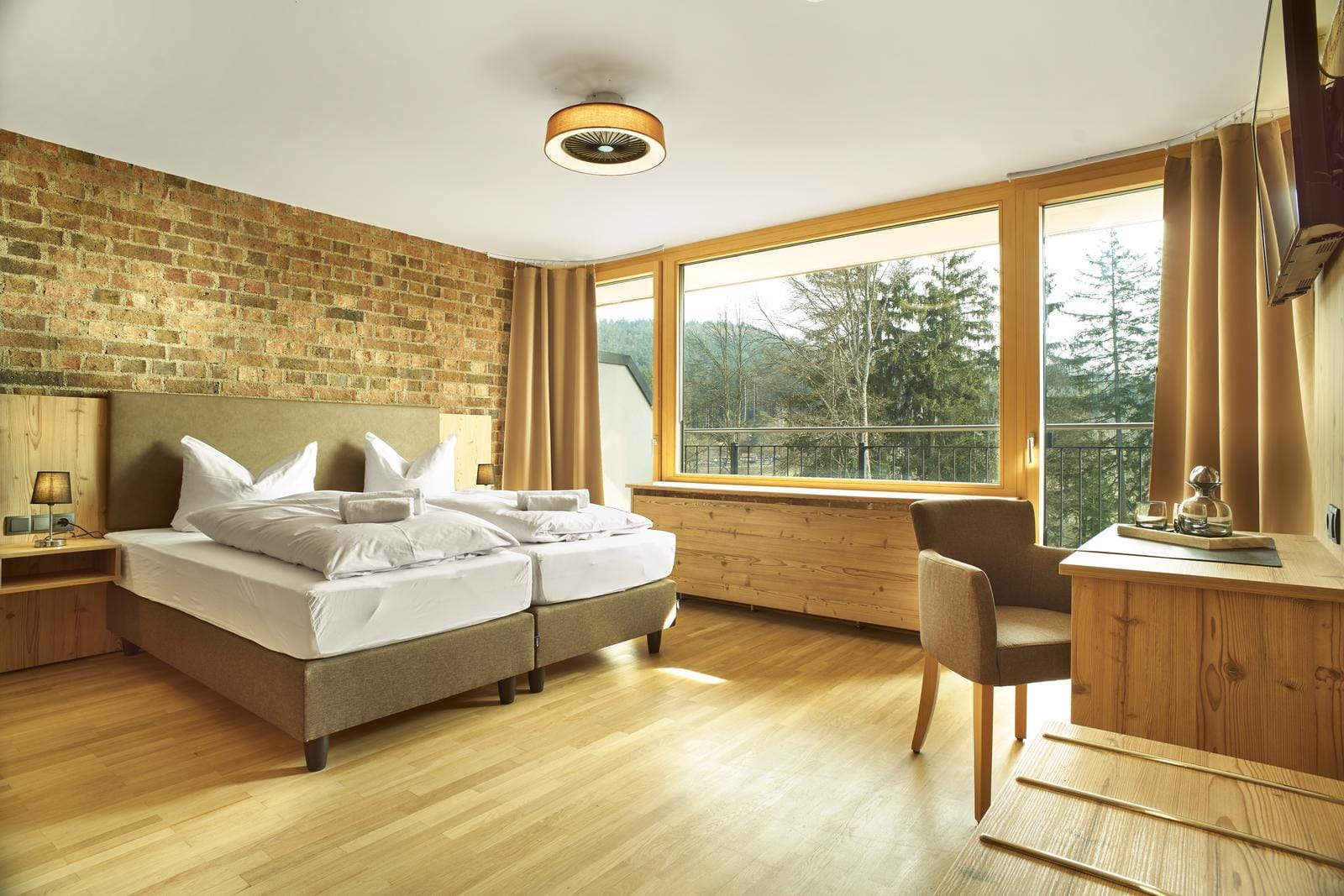 Modernes Hotelzimmer mit Naturholzmöbeln, Steinwand, großen Fenstern und Waldblick.