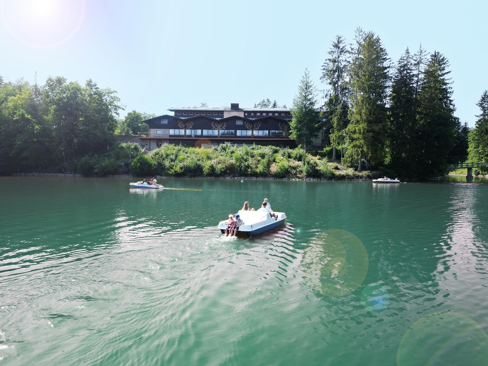 See mit Tretbooten und Wald, Hotel im Landhausstil im Hintergrund, sonniges Ambiente.