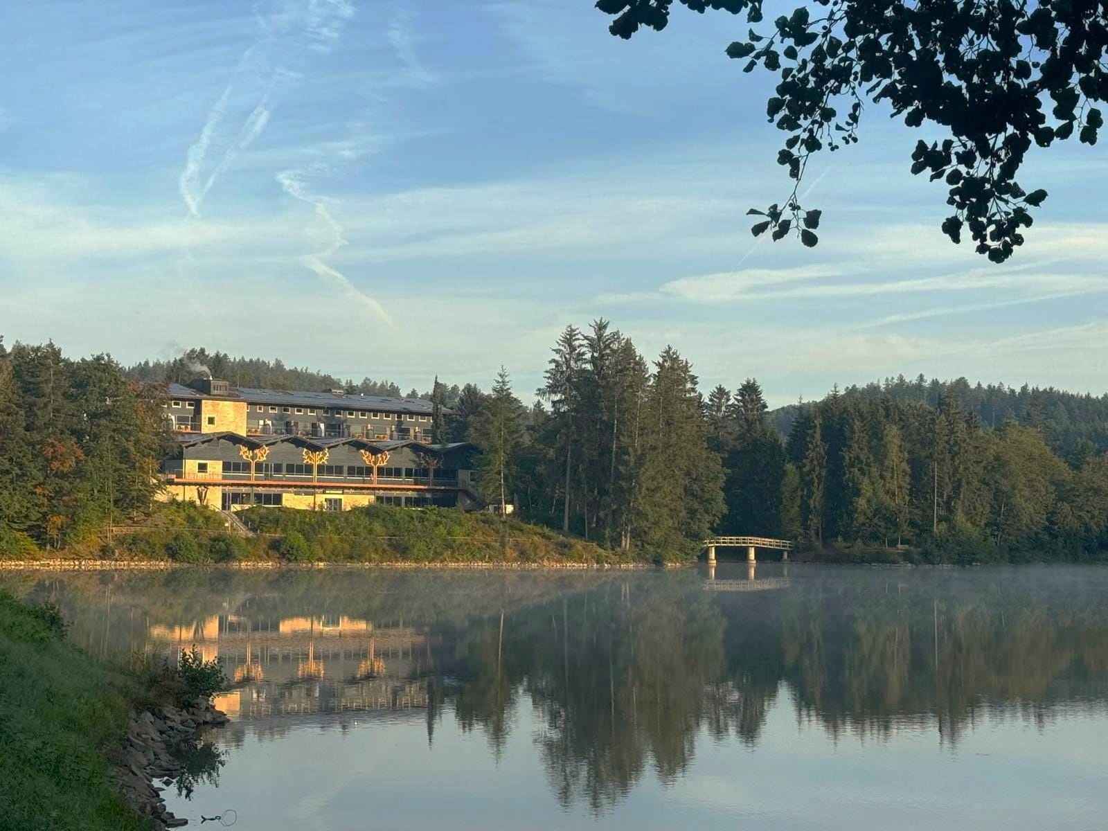 Hotel am Waldrand, am Seeufer gelegen mit spiegelnder Wasseroberfläche, ruhige Morgenstimmung.