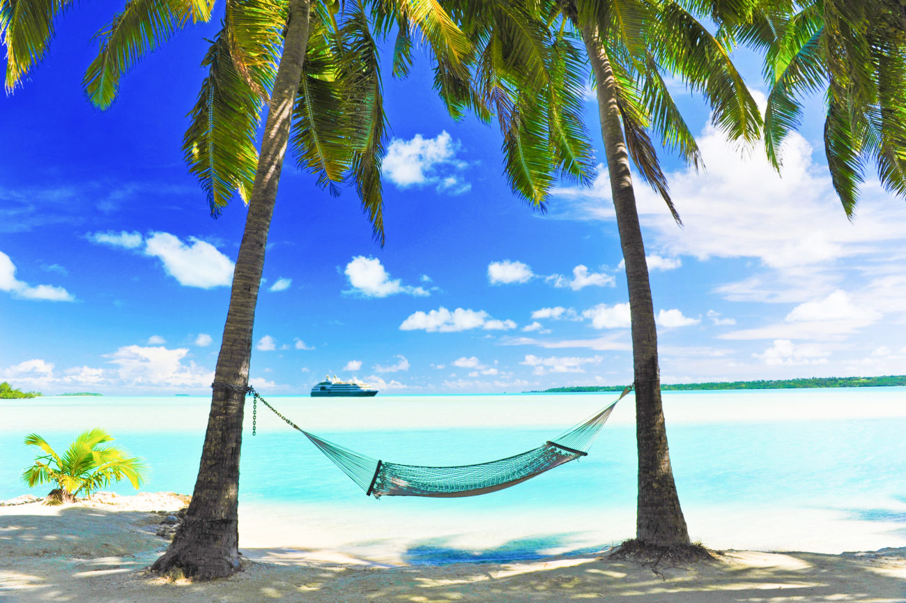 A cruise ship appears on the horizon of a tropical turquoise lagoon - with a hammock in semi silhouette, shaded by palm trees